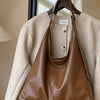 A brown PU leather slouchy hobo shoulder bag is displayed hanging on a hanger against a light beige coat.