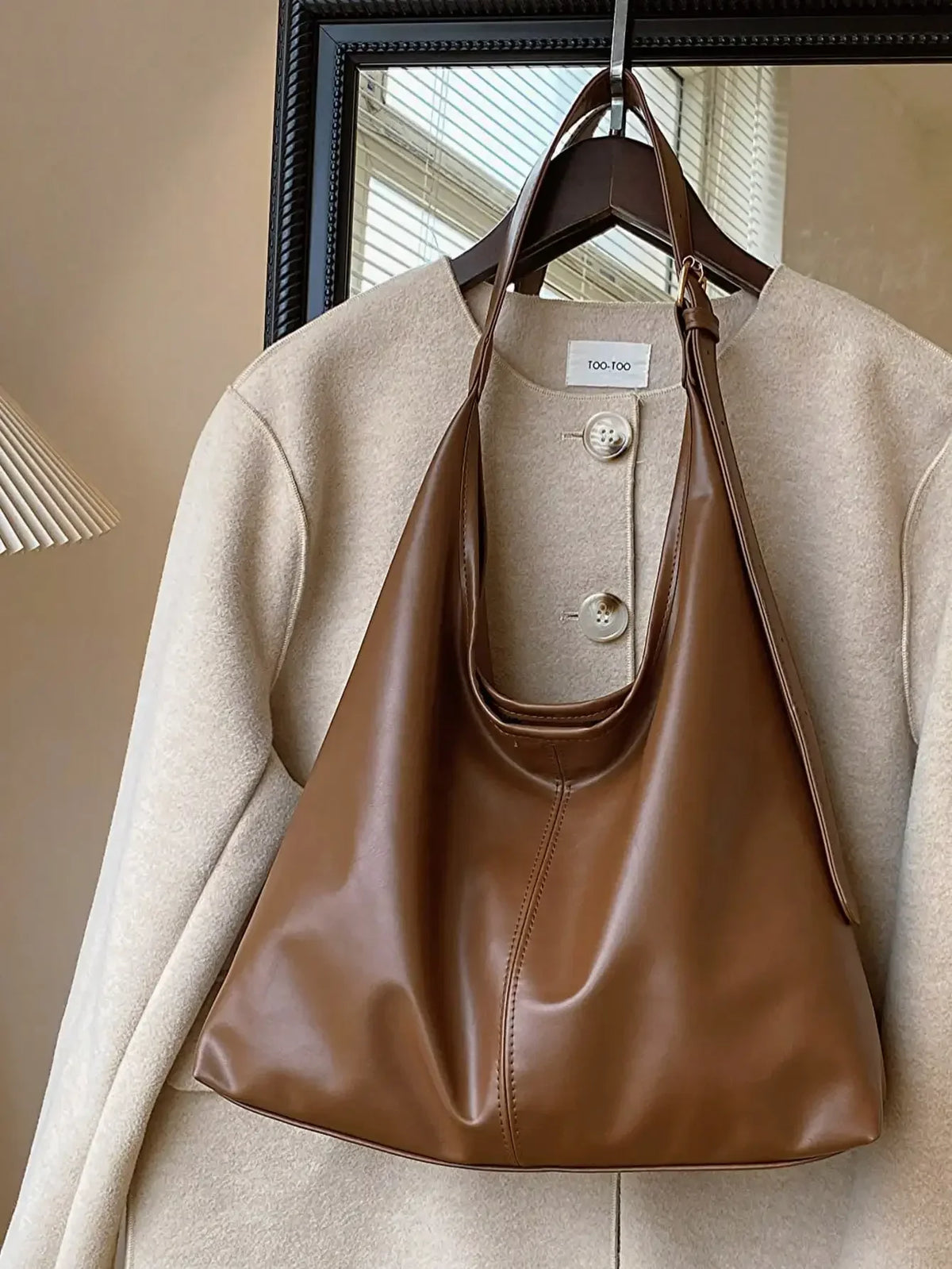 A brown PU leather slouchy hobo shoulder bag is displayed hanging on a hanger against a light beige coat.