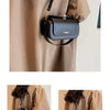 A black textured leather shoulder bag with a gold chain strap is shown being carried by a person wearing a beige trench coat.