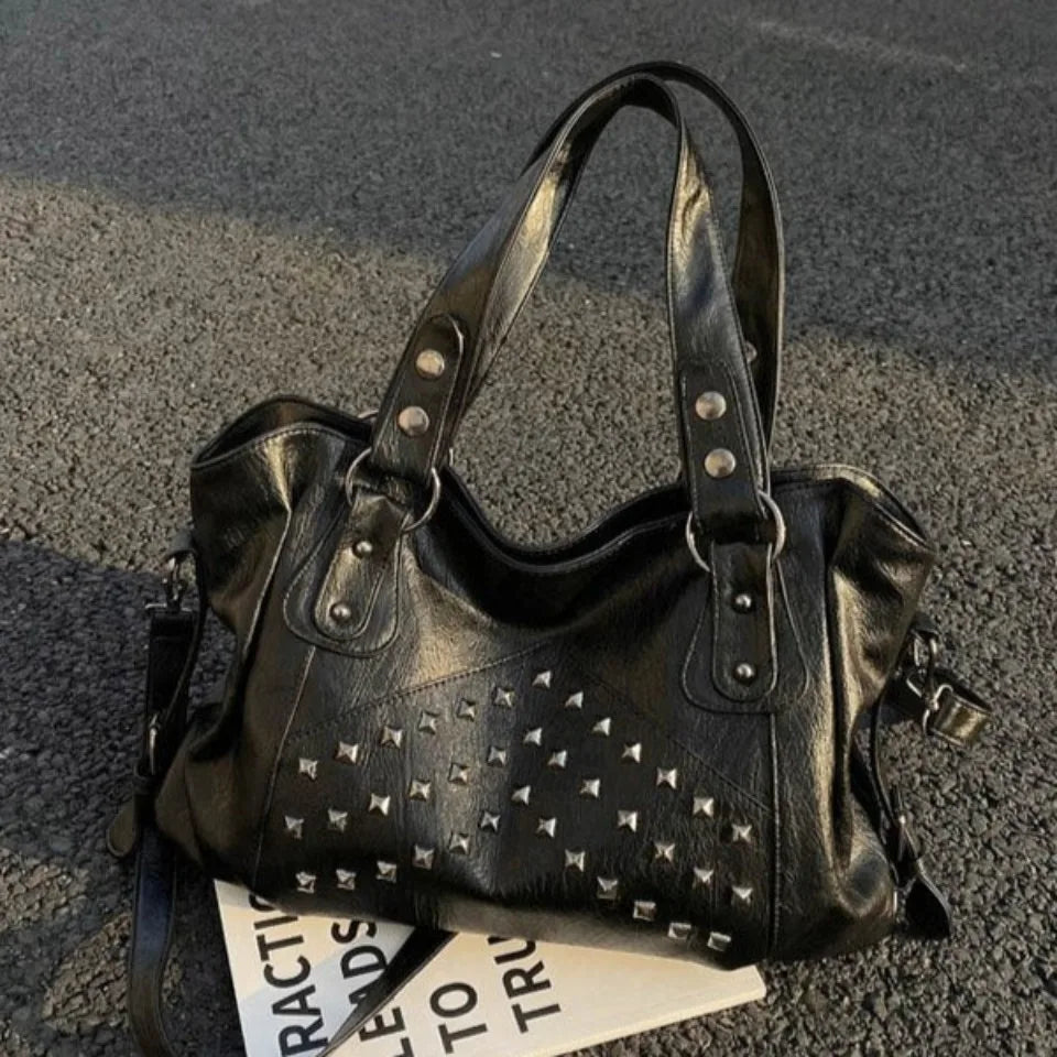A black punk rivet leather tote bag resting on an asphalt surface.