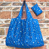 A bright blue Miffy folding shopping bag with a brown strap and a matching fabric square resting on a brick wall.