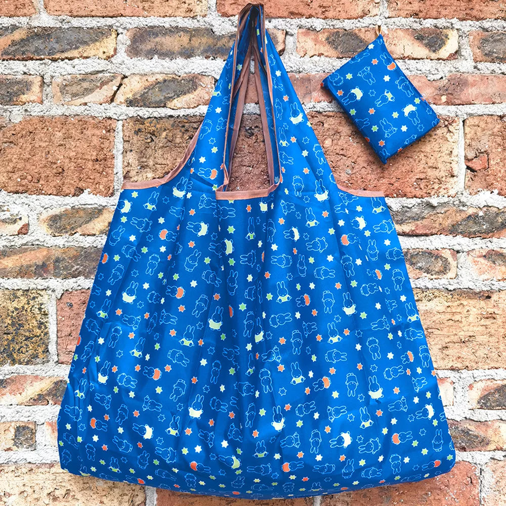 A bright blue Miffy folding shopping bag with a brown strap and a matching fabric square resting on a brick wall.