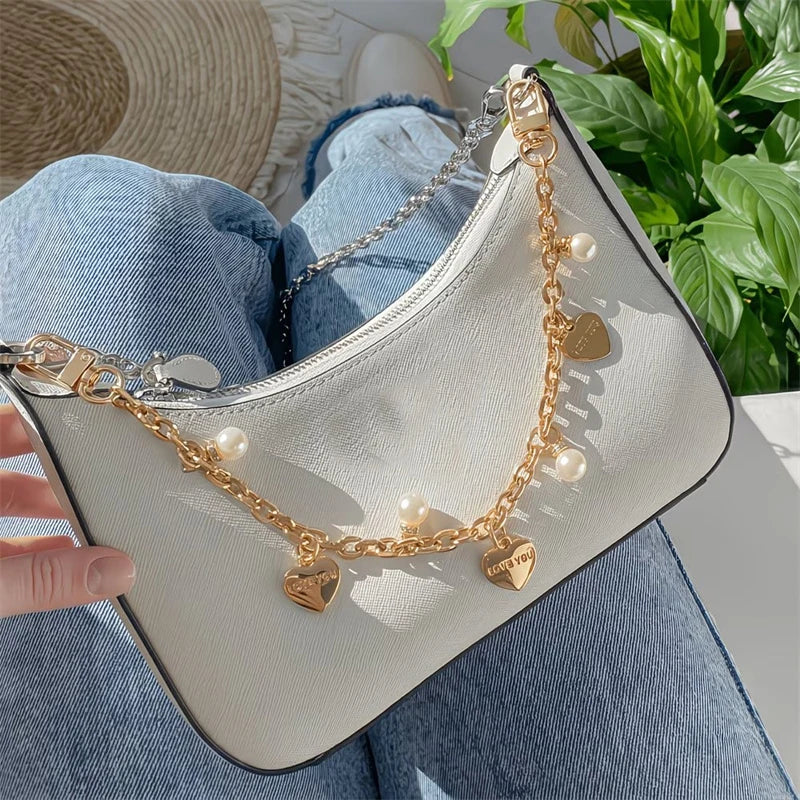 Helle beige Crossbody-Tasche mit goldener Kette, Perlen- und Herzanhängern, gehalten von einer Hand auf Denim.