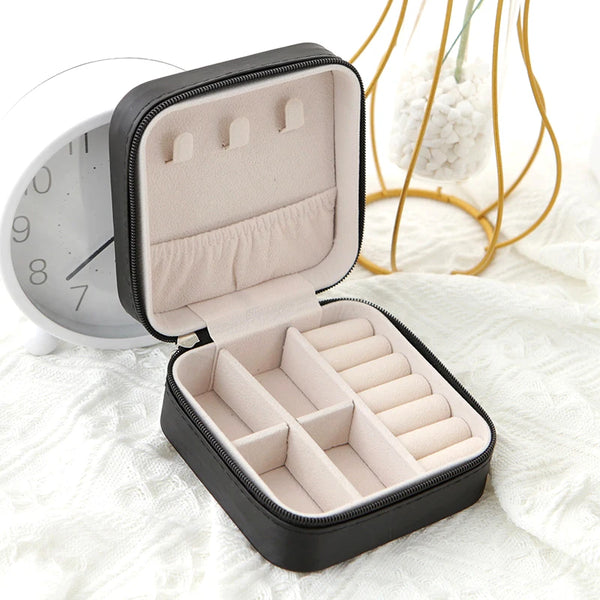 A black compact leather jewelry travel box with a light pink interior is shown open on a white surface.