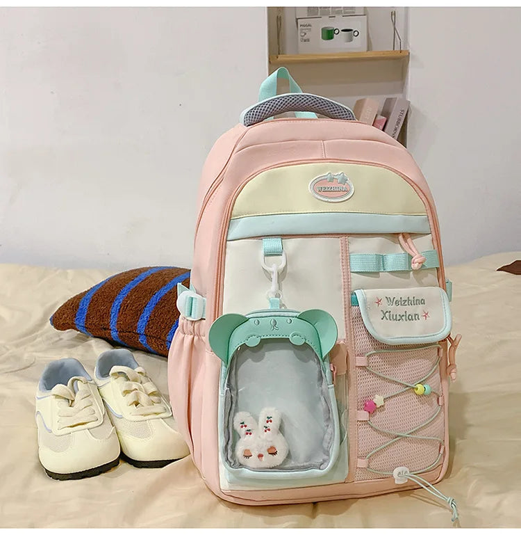 A pink and cream campus backpack with a light blue trim and a green accent, featuring a clear pocket and decorative elements.