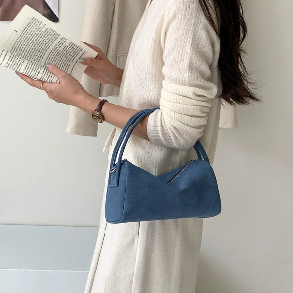 A woman holds a blue faux suede crossbody bag while wearing a cream-colored coat.