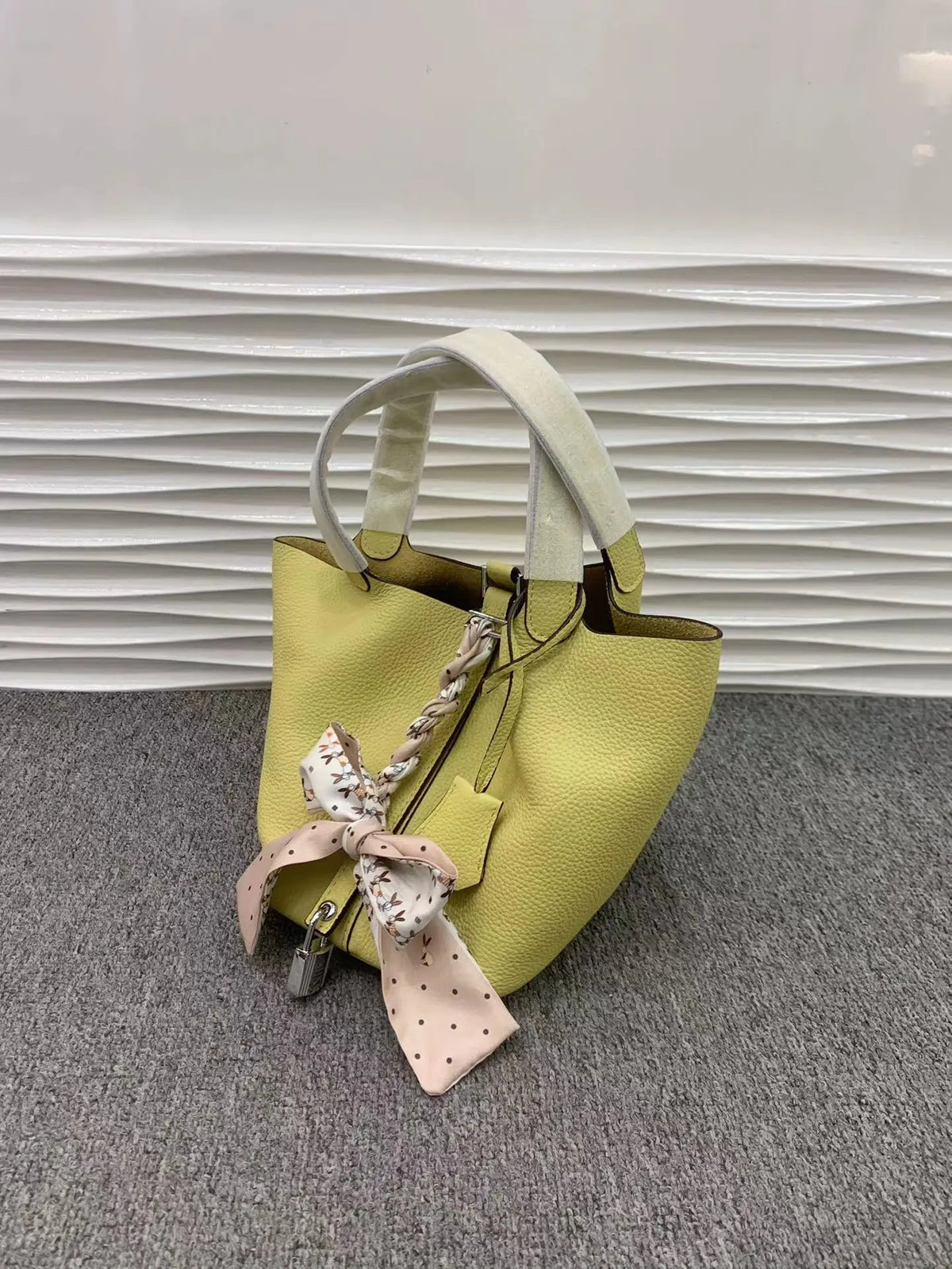 Helles gelbes Leder-Bucket-Bag mit weißem Riemen und rosa Blumenband, vor grauem Hintergrund.
