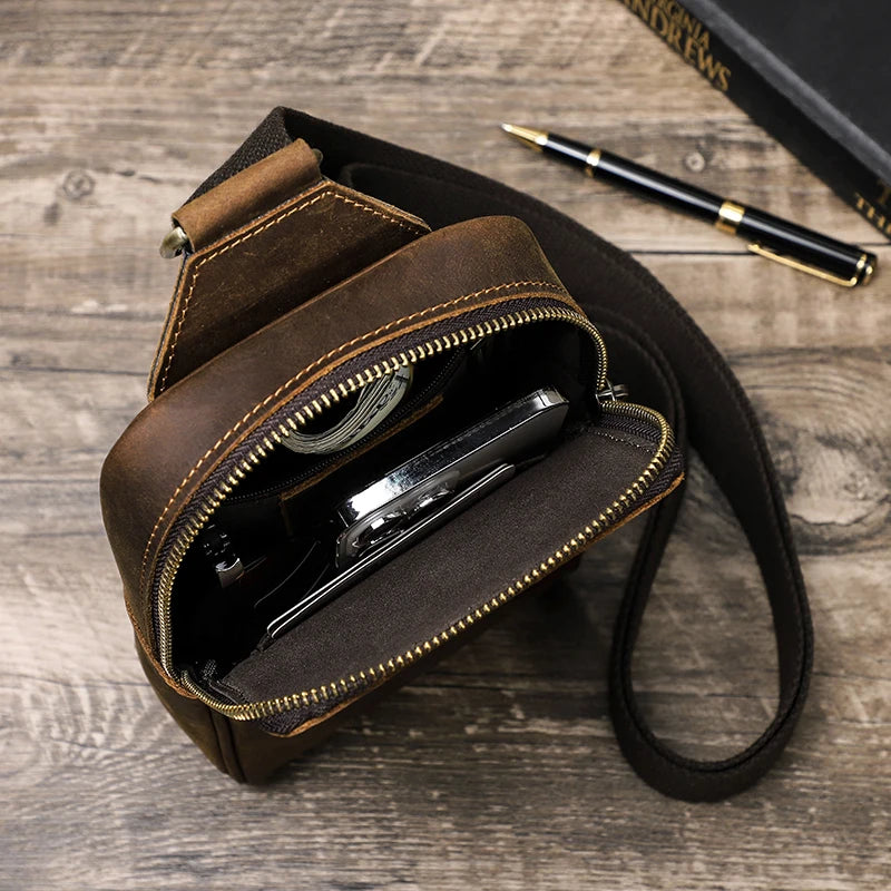 A brown genuine leather men's crossbody bag with a zippered compartment and a pen resting on a wooden surface.