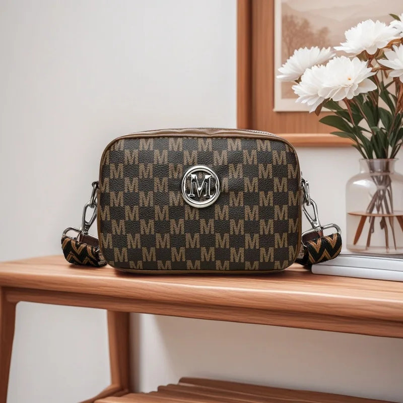 A brown patterned crossbody bag with silver hardware resting on a wooden surface next to a framed picture.