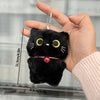 Una mano sostiene un colgante llavero de peluche de gato negro con una cadena de plata contra un fondo desenfocado.