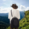 A person wearing a white shirt and hat is carrying a black EVA hard shell hat case while standing on a grassy hillside overlo