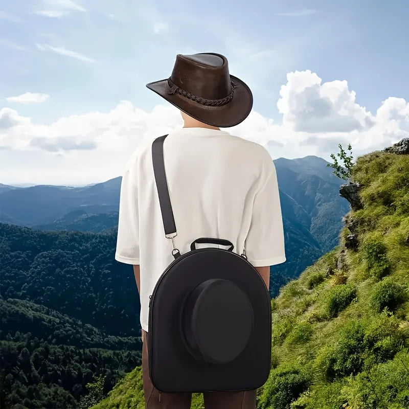 A person wearing a white shirt and hat is carrying a black EVA hard shell hat case while standing on a grassy hillside overlo