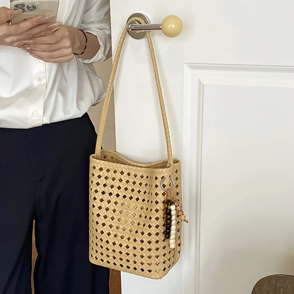 A light beige handwoven bucket bag with a woven pattern and wood bead tassel is hanging on a white wall.