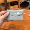 A hand holds a light gray genuine leather card holder with a gold stud and a white tag displaying a QR code and text.