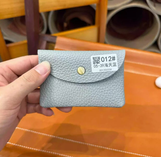 A hand holds a light gray genuine leather card holder with a gold stud and a white tag displaying a QR code and text.