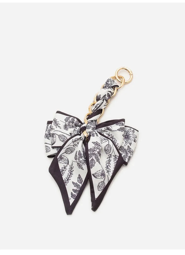 A black and white floral patterned bow made of fabric is attached to a gold chain strap extension for a handbag.