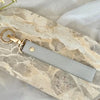 A light gray leather wristlet keychain with a gold-toned clasp rests on a marbled stone surface.