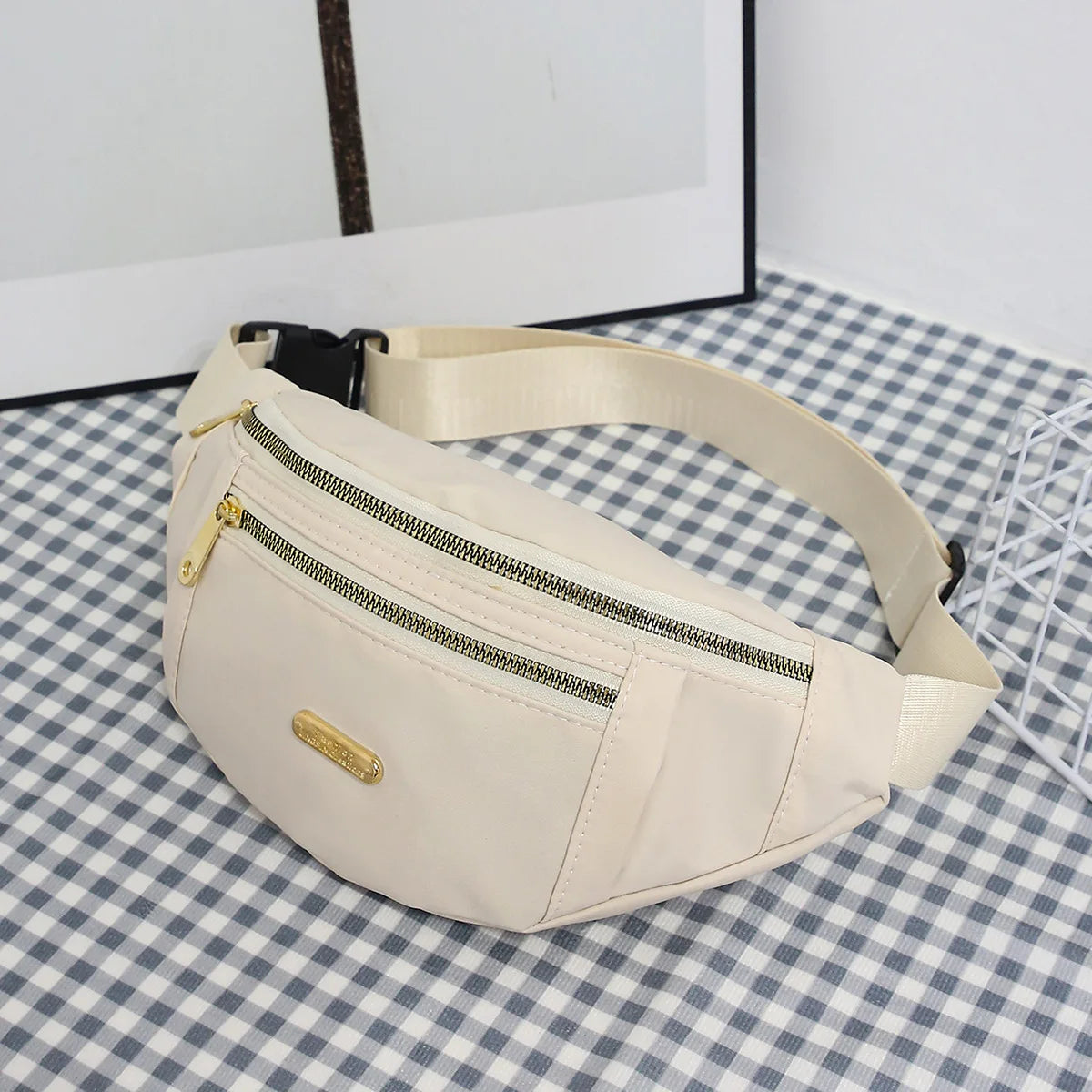 A beige lightweight nylon crossbody fanny pack with a zippered pocket is displayed on a checkered fabric surface.