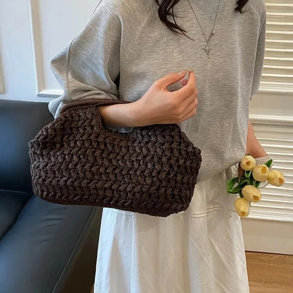 A woman holds a dark brown straw woven tote handbag against a light gray shirt and white skirt.
