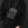 A man wearing a black jacket is holding an olive green Oxford chest bag with a strap.