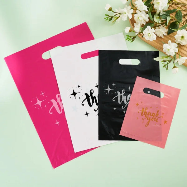 Three reusable plastic thank you bags in pink, white, and black are displayed on a light green surface.
