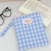 A light blue plaid cotton tablet pouch with a pink tag rests on a white surface next to glasses and a book.