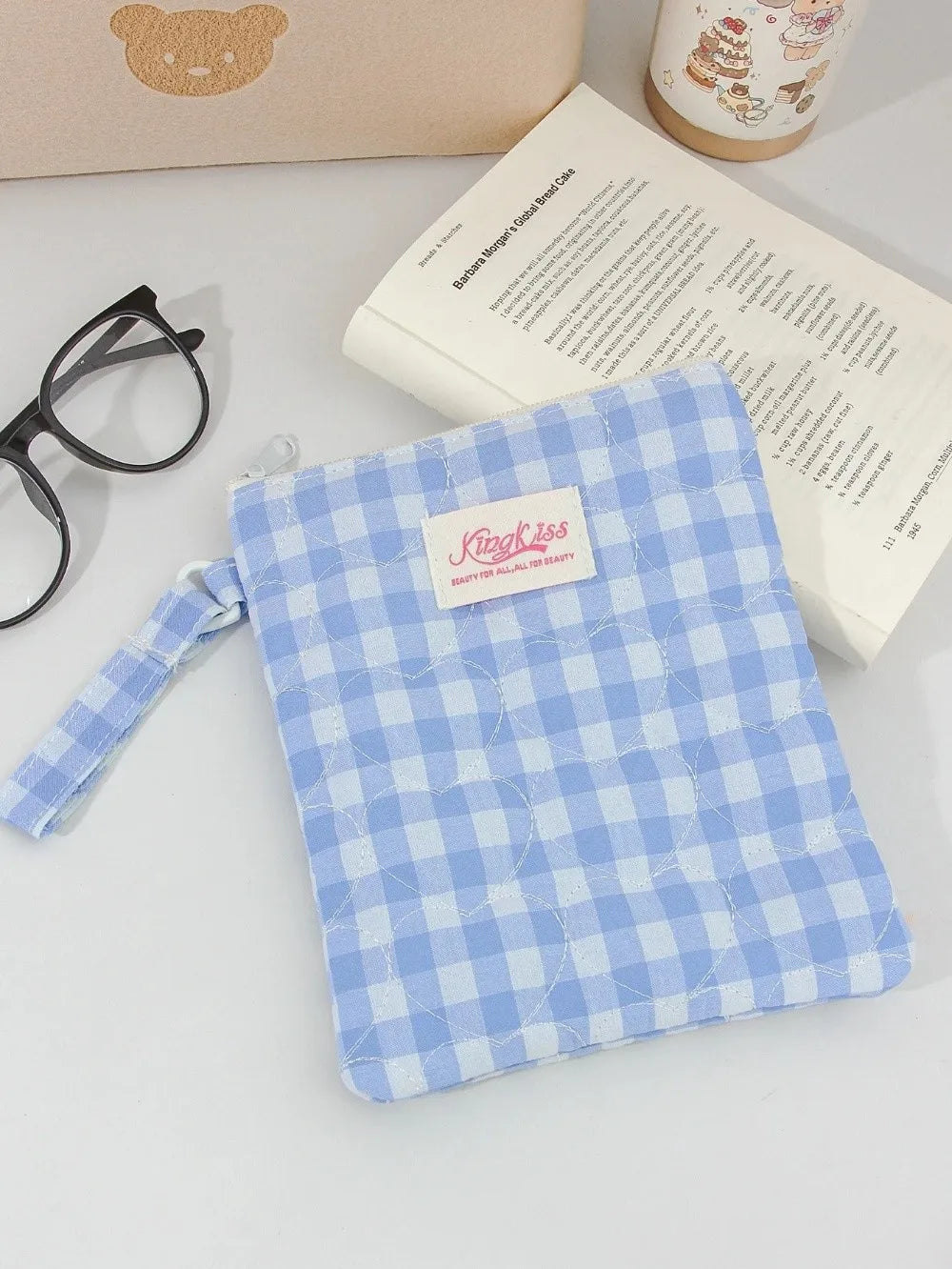 A light blue plaid cotton tablet pouch with a pink tag rests on a white surface next to glasses and a book.