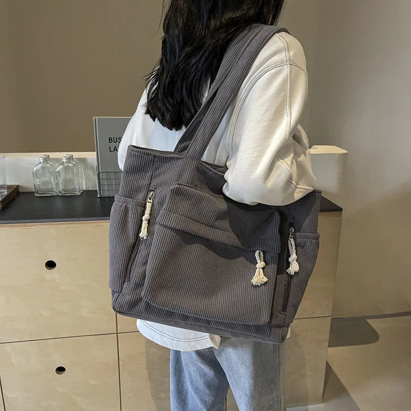 A person is holding a gray corduroy shoulder bag with a zipper on their back in a light indoor setting.