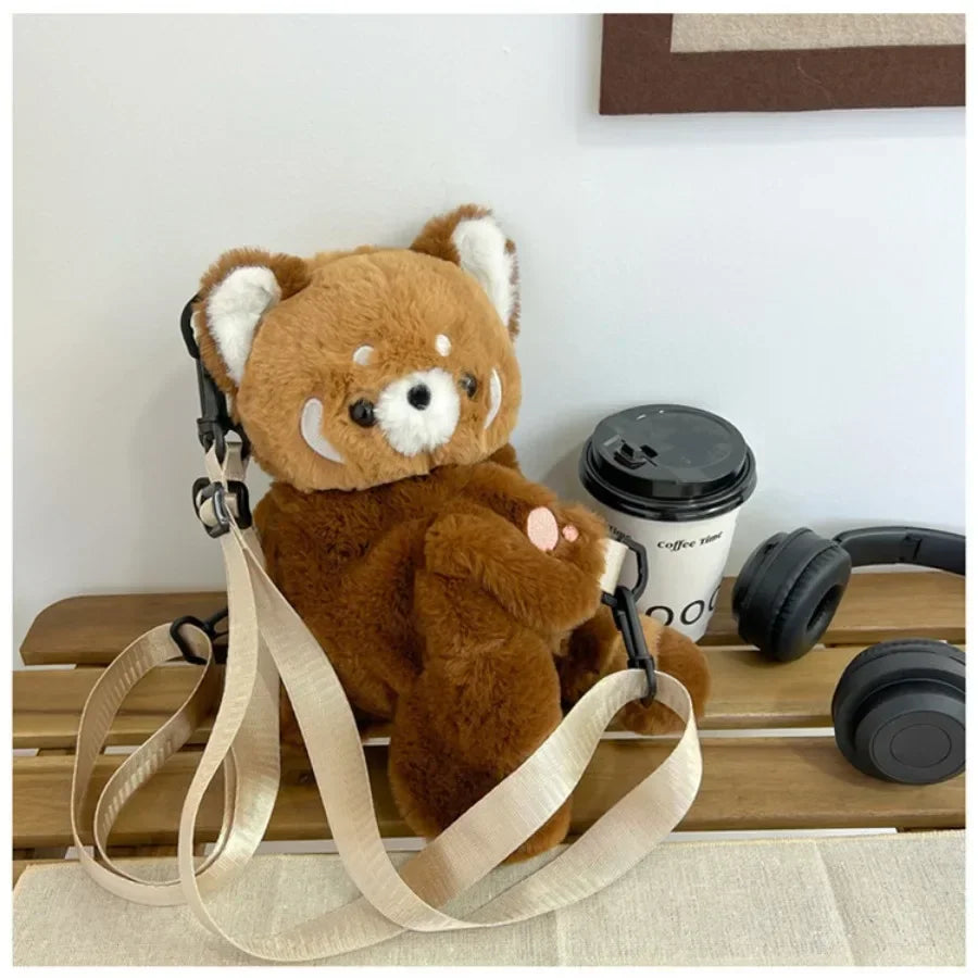 A brown red panda plush toy is displayed next to a black camera lens and leather straps on a wooden surface.