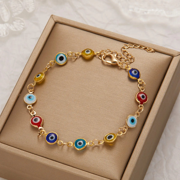 A gold charm bracelet featuring blue, red, and yellow evil eye beads displayed in a light brown box.