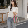 A woman wearing a cream knit sweater and jeans holds a pink and green striped bucket tote bag.