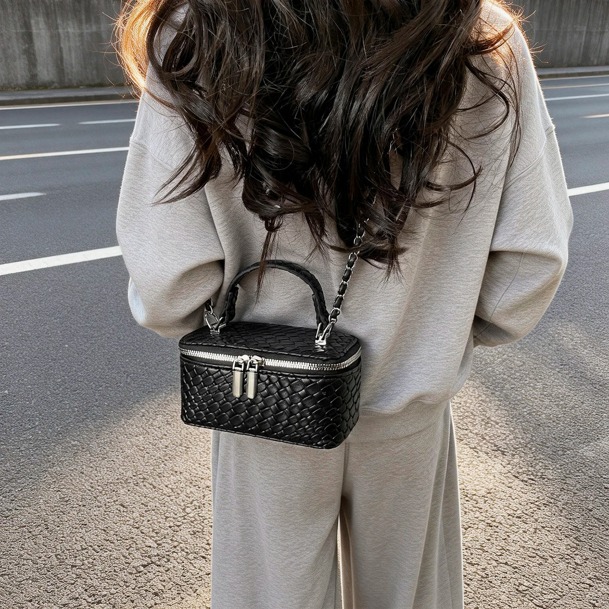 A woman is holding a black woven crossbody bag with a silver chain strap against a gray background.