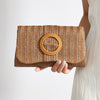 A hand holds a woven straw envelope clutch with a circular detail against a light background.