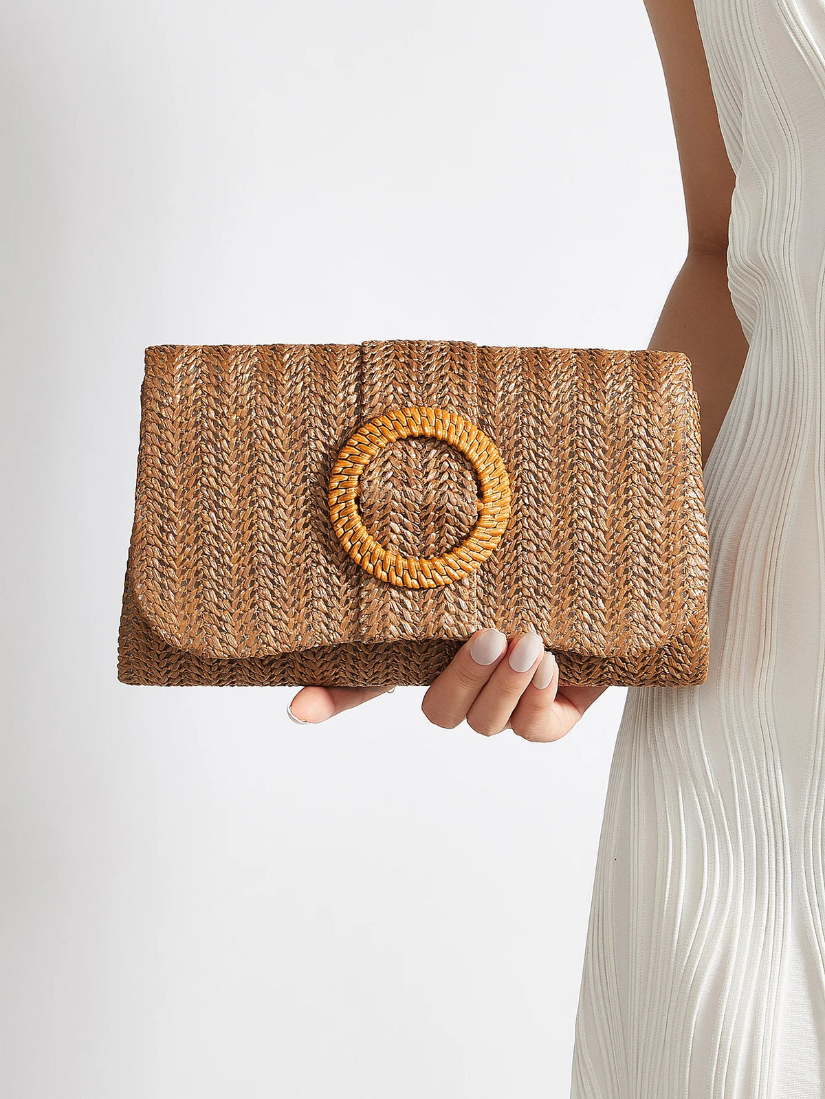 A hand holds a woven straw envelope clutch with a circular detail against a light background.