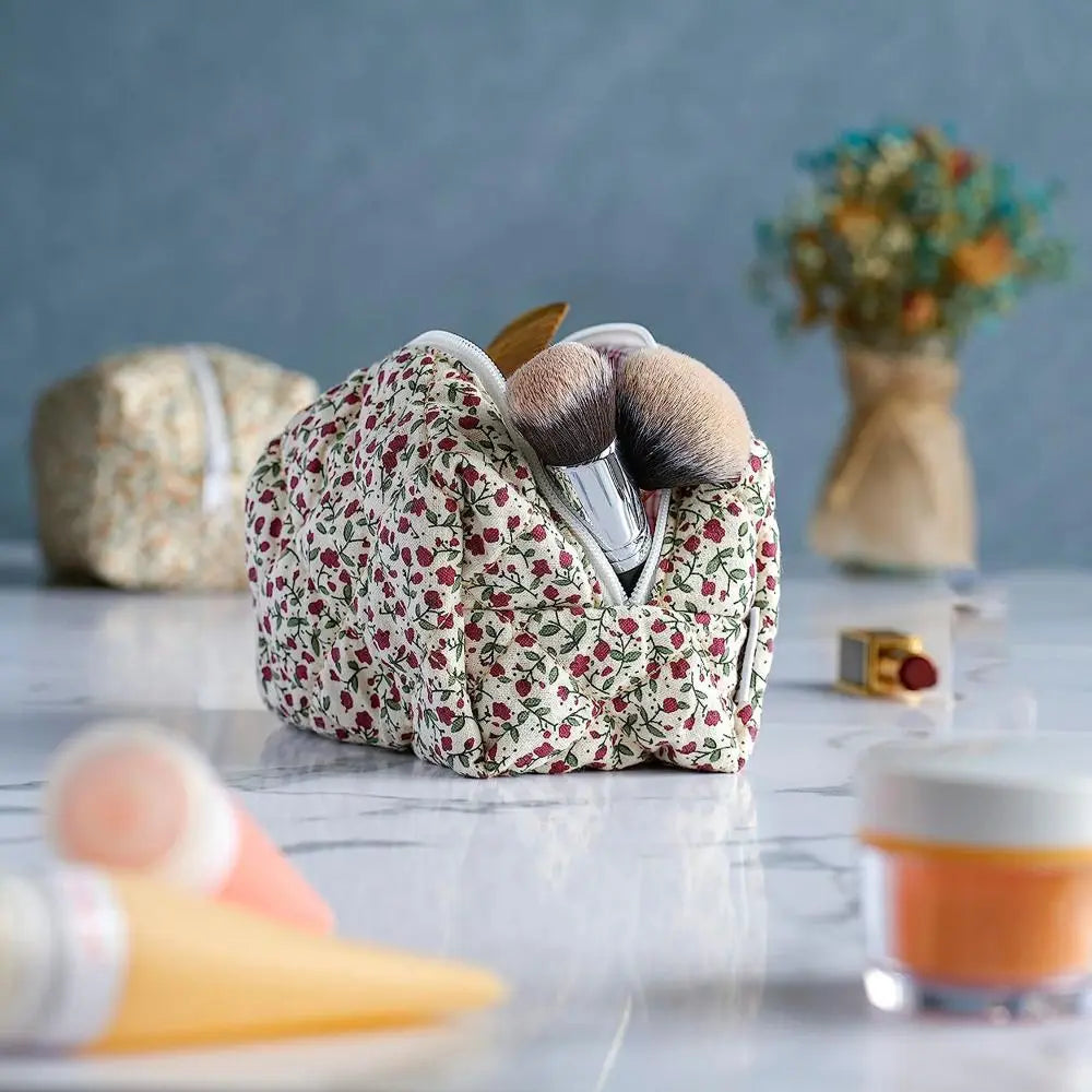 Bolsa de maquillaje acolchada con cremallera floral llena de brochas y un pequeño jarrón de flores sobre una superficie blanc