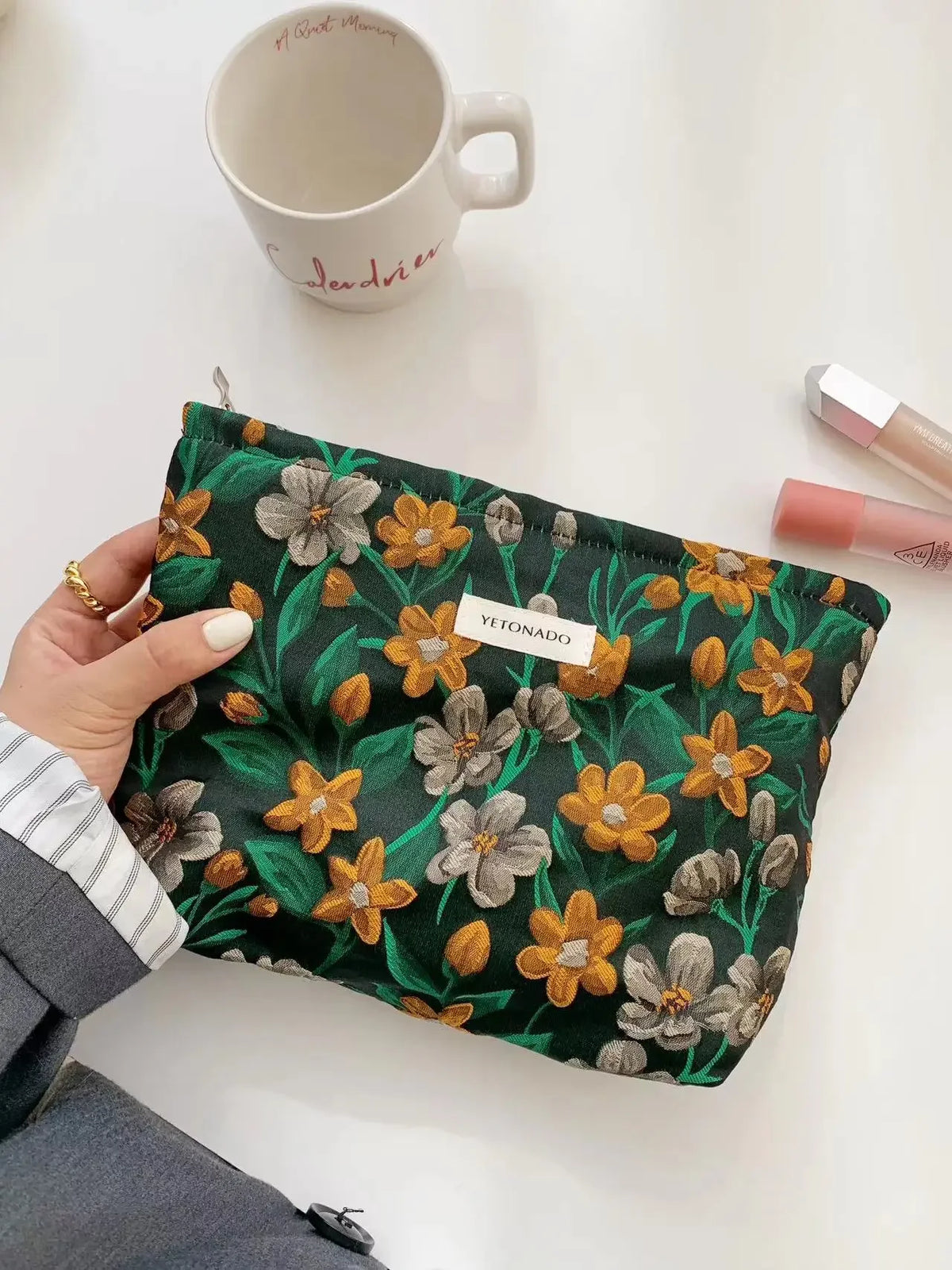 A dark green cosmetic pouch with a floral pattern rests on a white surface next to a ceramic mug.