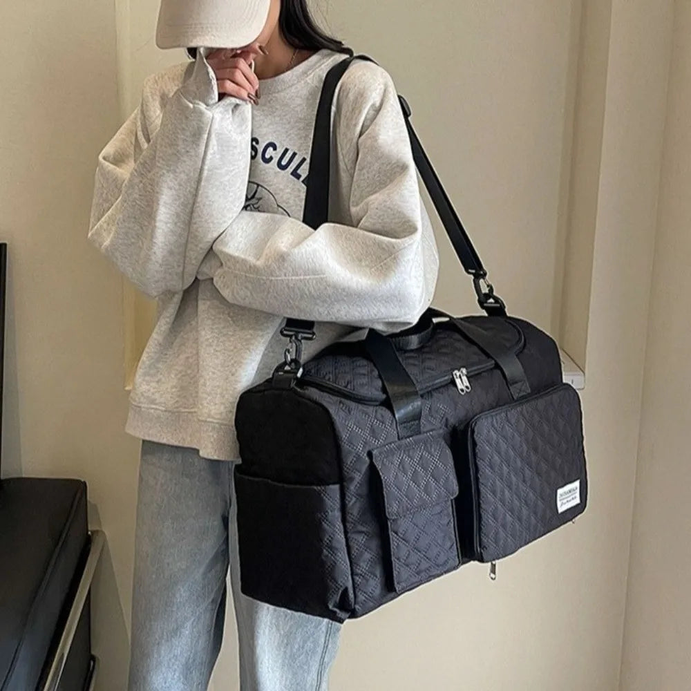A person wearing a light gray sweatshirt holds a black quilted travel duffle bag over their shoulder.