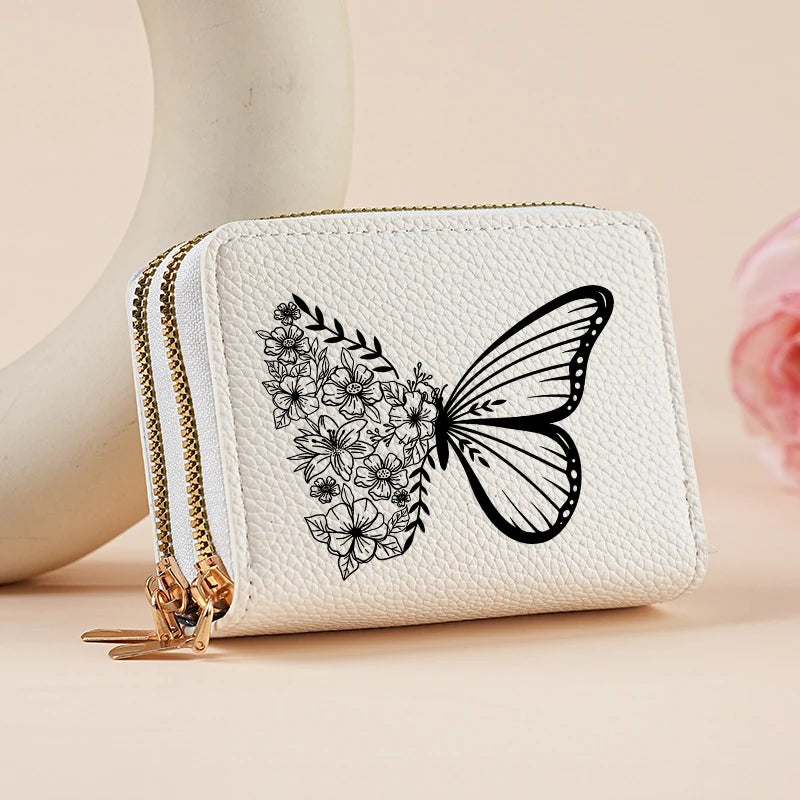 A white zippered wallet featuring a black floral butterfly print, resting on a light pink surface.