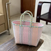 A hand woven pink and yellow striped PVC vegetable basket tote bag sitting on a dark wooden surface.