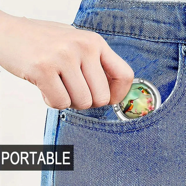 A hand is holding a small, oval-shaped bag hook with a bird design against a denim fabric.