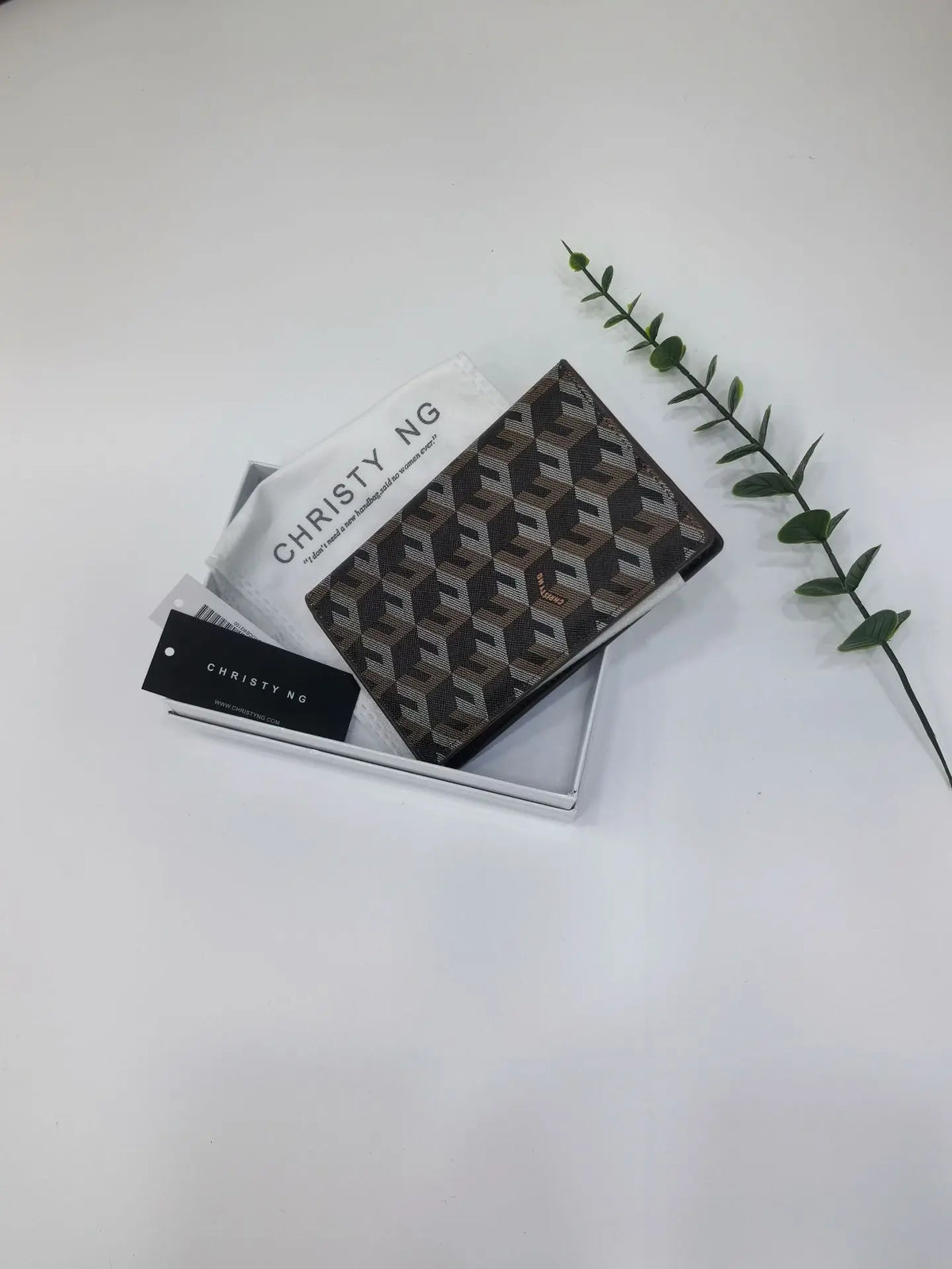 A brown geometric print passport wallet is displayed next to a white card and a green leafy branch.