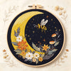 A dark fabric embroidery featuring a yellow crescent moon and floral elements, with a bee flying near the moon.