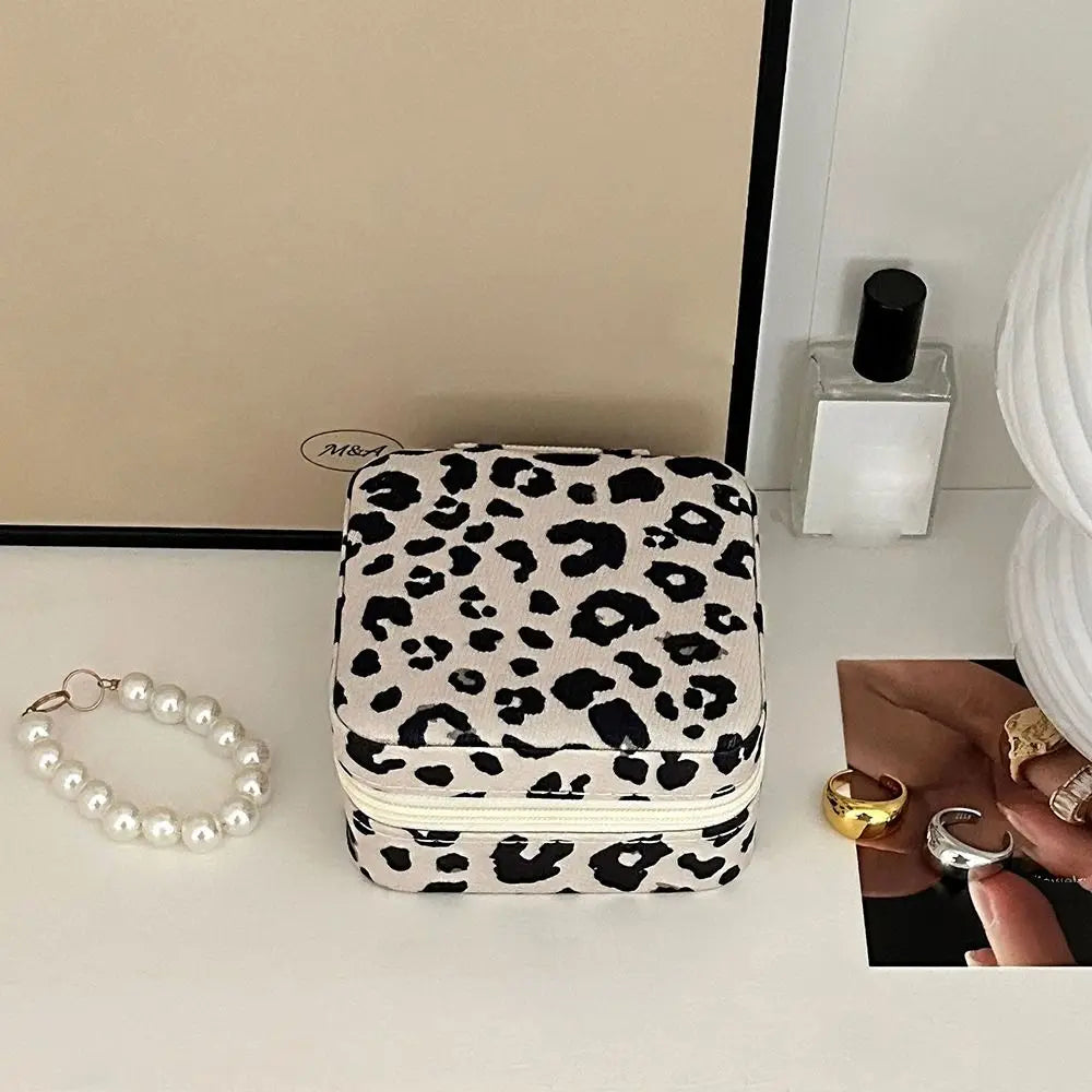 A leopard print PU leather jewelry storage box is displayed on a white surface next to some gold rings and a white candle.