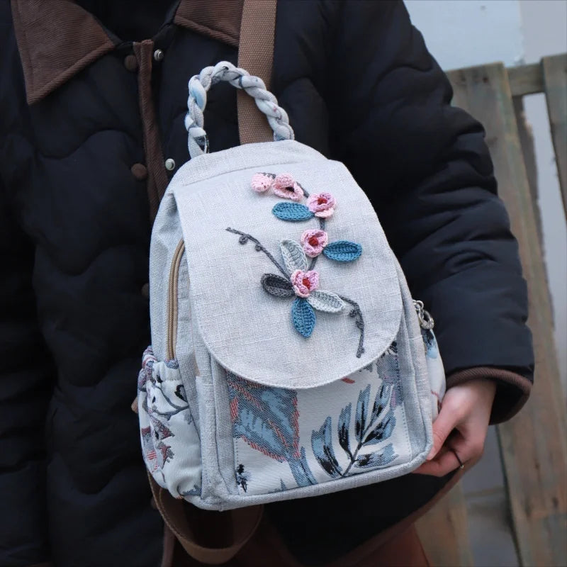 A light gray linen backpack featuring delicate pink and blue 3D floral embroidery.
