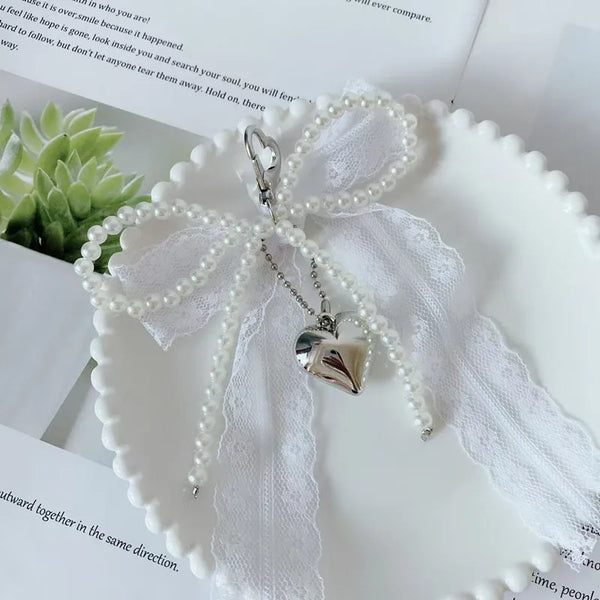 A white lace bow keychain with pearl strands and a silver heart pendant resting on a white surface.