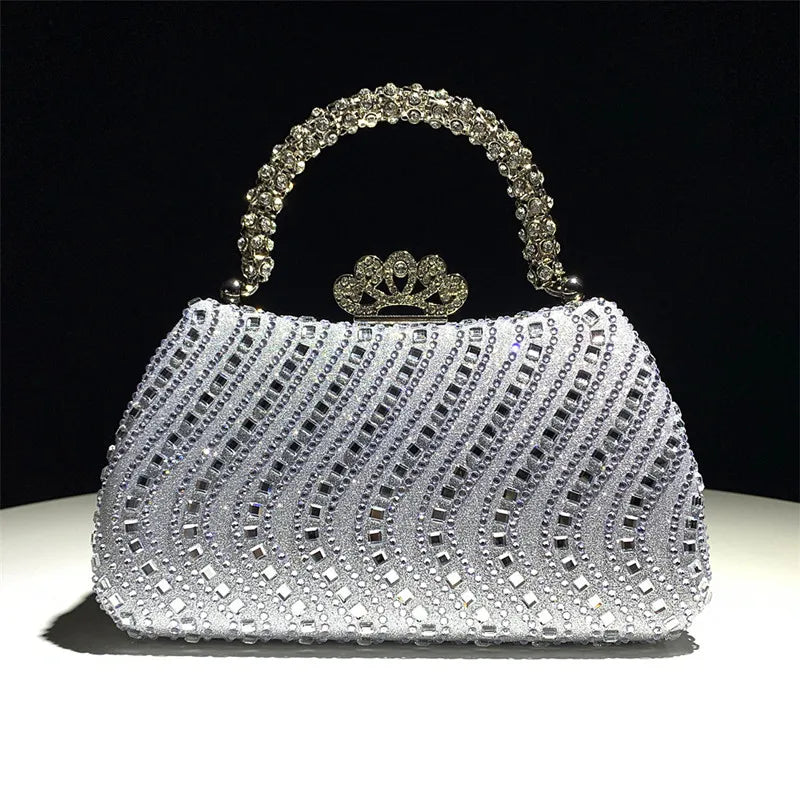 A silver crystal-embellished evening clutch bag with a crown-shaped clasp and chain strap, resting on a white surface.