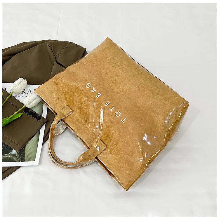 A tan transparent waterproof PVC tote bag is shown next to a dark olive green fabric item and a white flower.
