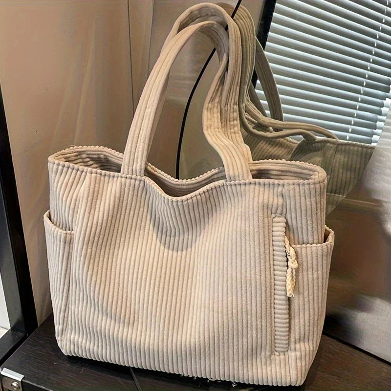 A beige casual corduroy handbag with vertical ribbing is displayed in front of a window blind.