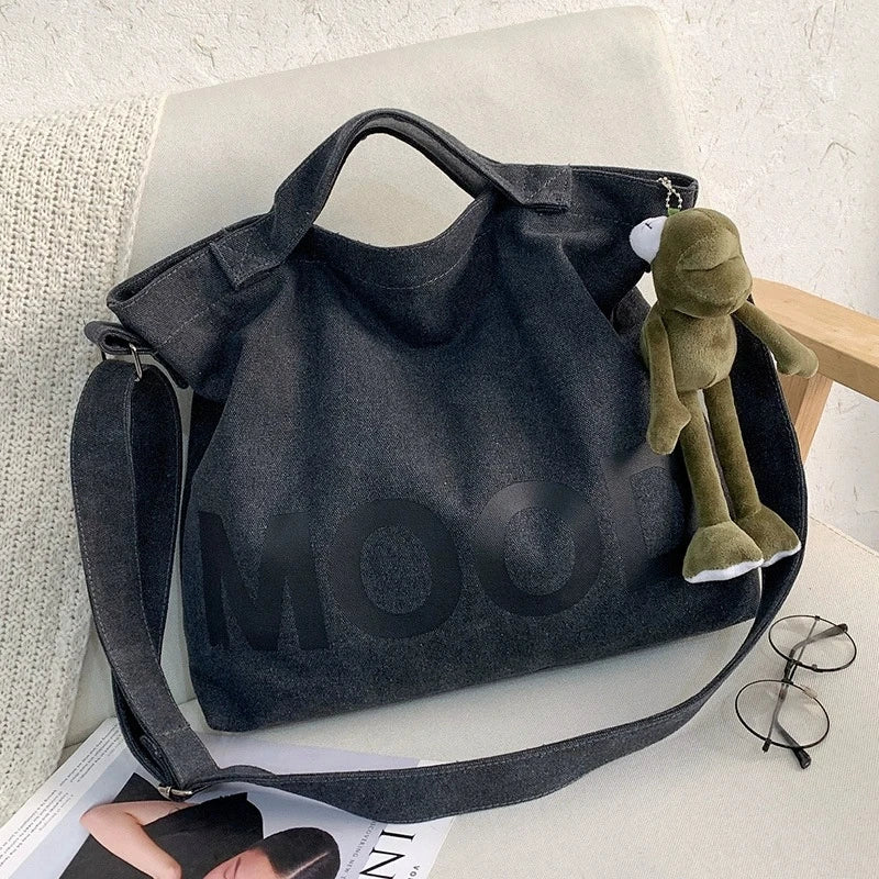 A dark gray canvas tote bag featuring a large letter design and a small green stuffed animal resting on it.