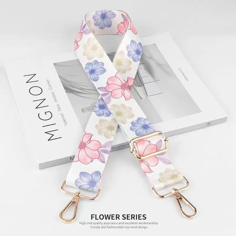 A white floral patterned nylon bag strap with a gold buckle is displayed on a wooden surface.